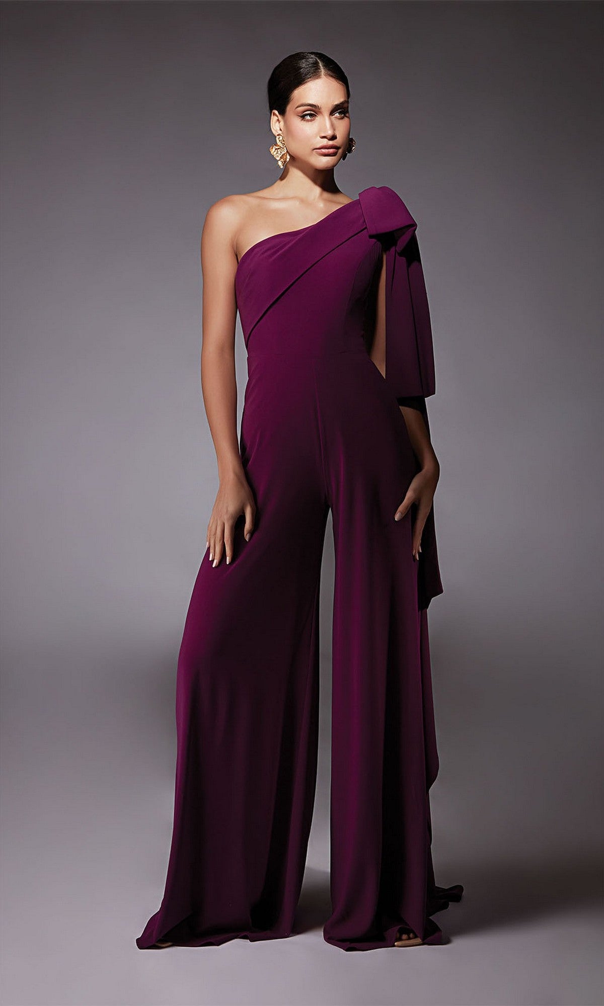 A person wearing the Alyce 70065 long formal jumpsuit in deep purple, featuring one-shoulder styling, wide legs, and a large bow detail—an elegant cocktail dress alternative—stands against a plain gray background.